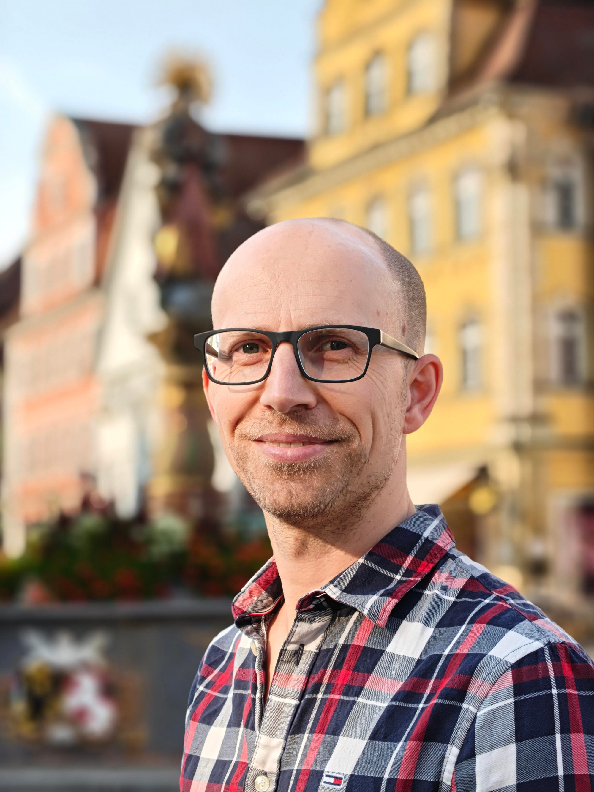 Simon Hegener looking into the camera smiling, standing in front of a fountain in a small town